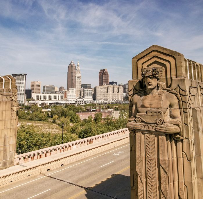 Art Deco statues on a bridge overlooking a city skyline with skyscrapers and clear blue skies.