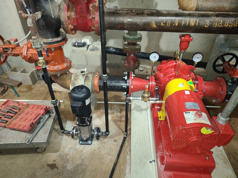 Industrial pump system with red and black components, pipes, gauges, and control valves in a mechanical room.