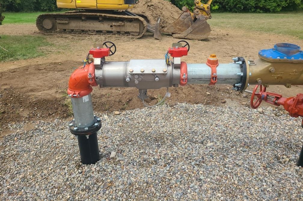 A pipeline with red valves and a pressure gauge is installed on a gravel surface, with construction equipment in the background.